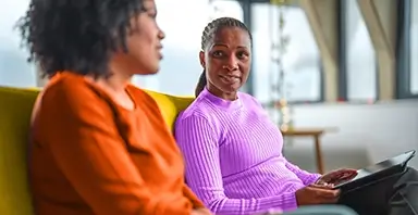 Two women sit on a yellow couch indoors, having a conversation. One woman holds a tablet while listening to the other, who is speaking.