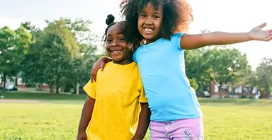 Two young children stand outdoors on grass, smiling at the camera. One child has an arm around the other, and the other extends an arm out to the side. Trees and sunlight are in the background.