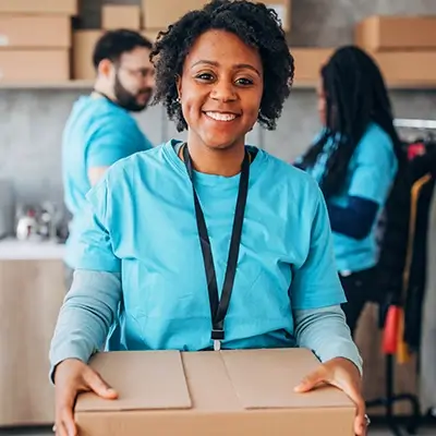 A woman in a blue shirt holds a cardboard box and smiles at the camera, with two people working in the background.