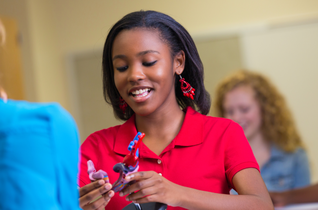 A young woman in a red shirt and earrings examines an anatomical model in a classroom, with another student blurred in the background.