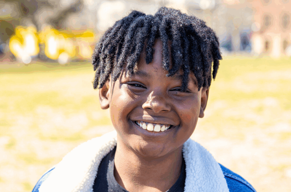 Smiling person with short, textured hair standing outdoors in bright sunlight, wearing a denim jacket with a white fleece collar.