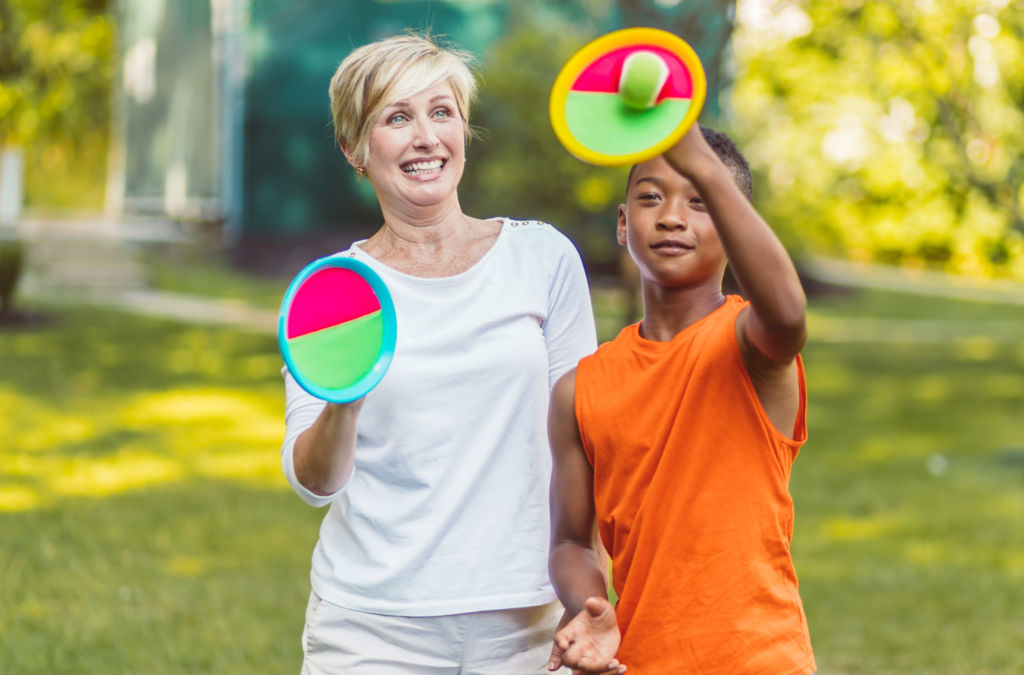An adult and a child play catch with velcro paddle and ball toys outside on a sunny day.
