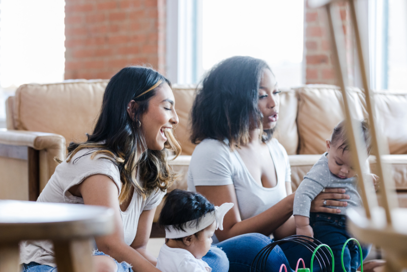Two women sit on a couch with two babies in a living room, one woman holding a baby and the other next to a baby with a headband.