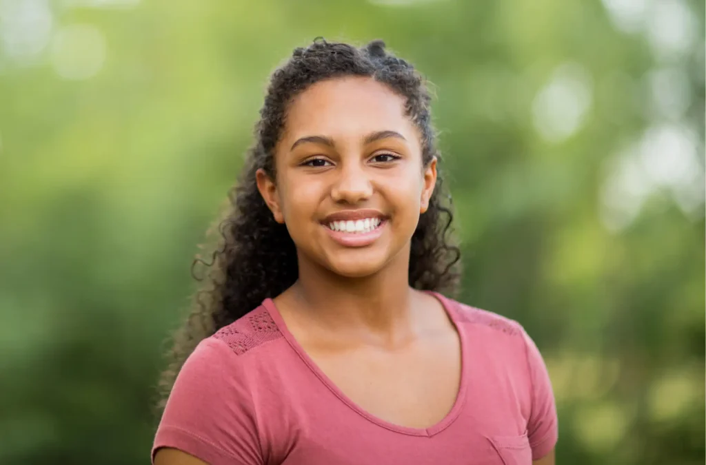A young woman with curly hair, wearing a pink t-shirt, smiles at the camera outdoors with a blurred green background.