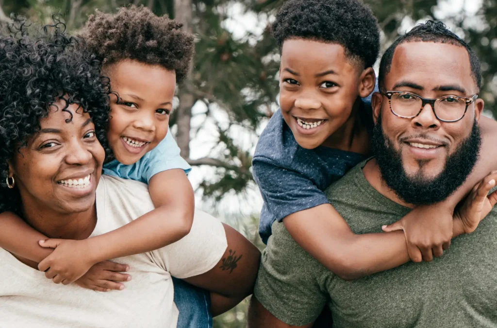 A family of four poses outdoors, with two children smiling and riding on the backs of two adults, all looking at the camera.
