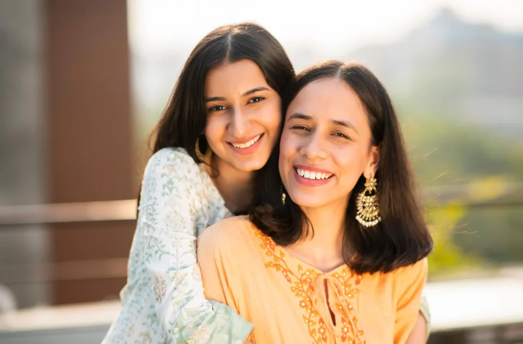 Two women smile outdoors, one standing behind the other and hugging her. Both are wearing traditional attire and earrings, with greenery and sunlight in the background.