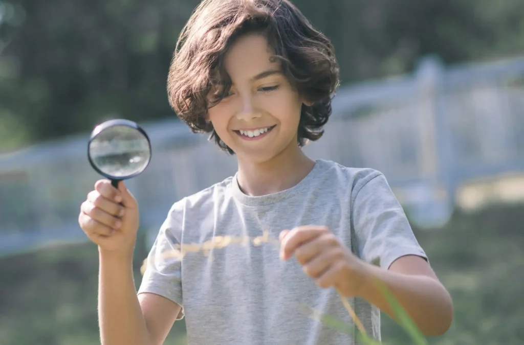 Smiling child holding a magnifying glass examines a plant outdoors on a sunny day.