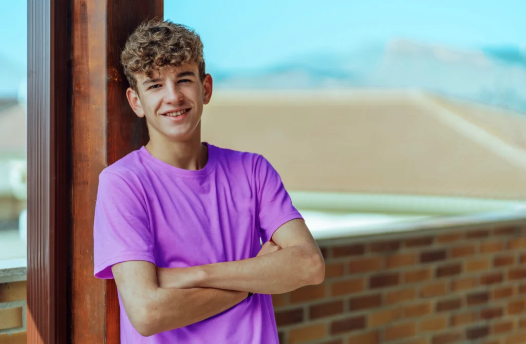 Teenager with curly hair wearing a purple t-shirt stands outside, leaning against a wooden post with arms crossed, smiling at the camera. A brick wall and rooftops are in the background.