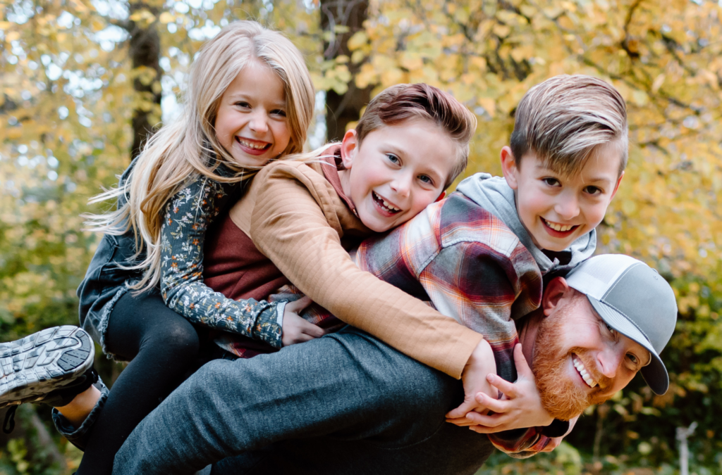 An adult man in a hat holds three smiling children on his back outdoors, surrounded by autumn foliage.