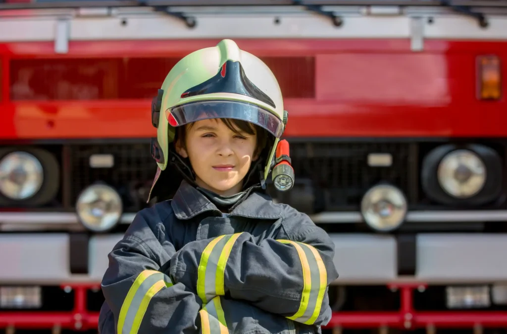 A child wearing a firefighter helmet and uniform stands with arms crossed in front of a red fire truck.