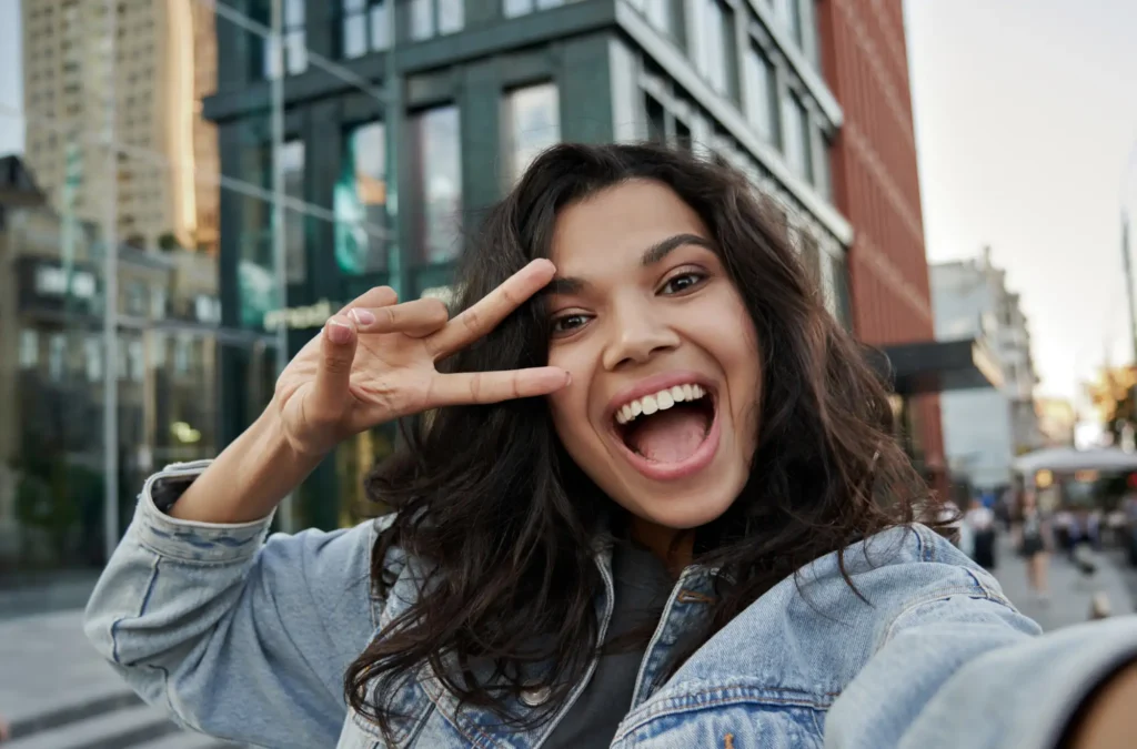 A young woman smiles and makes a peace sign while taking a selfie outdoors in an urban area with tall buildings in the background.