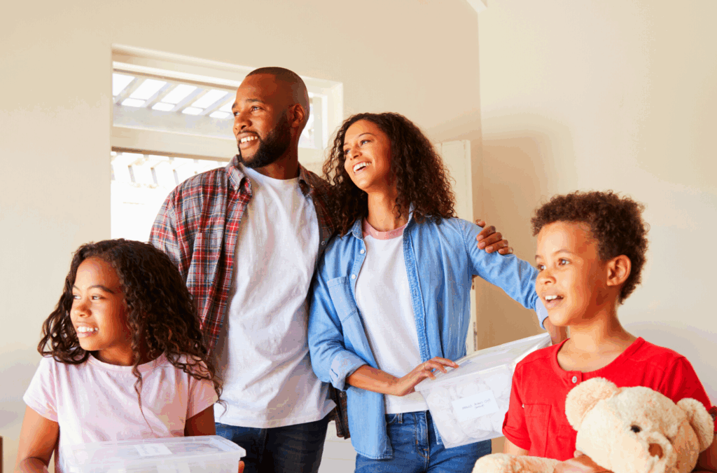 A family of four stands indoors, smiling and holding moving boxes and a teddy bear, suggesting they are moving into a new home.