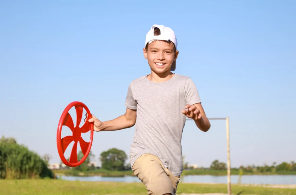 A boy wearing a white cap and gray t-shirt is running outside on grass while holding a red flying disc. Blue sky and trees are visible in the background.