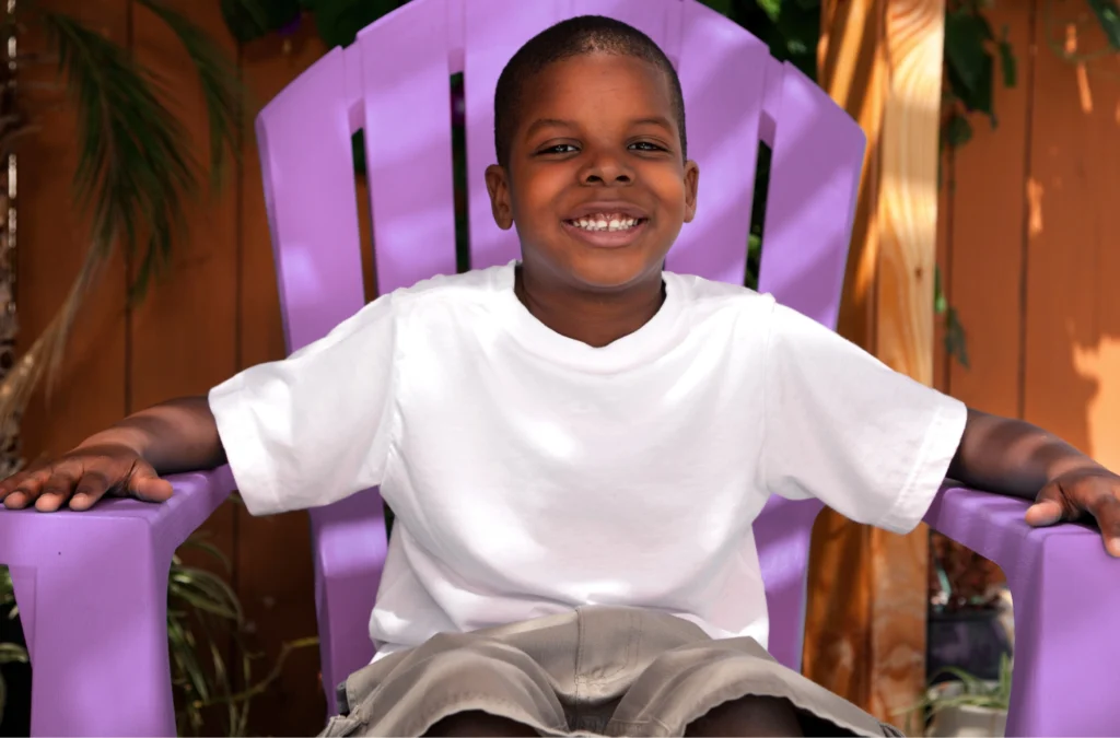 A young boy in a white t-shirt and khaki shorts sits smiling on a purple outdoor chair, with a wooden fence and plants in the background.