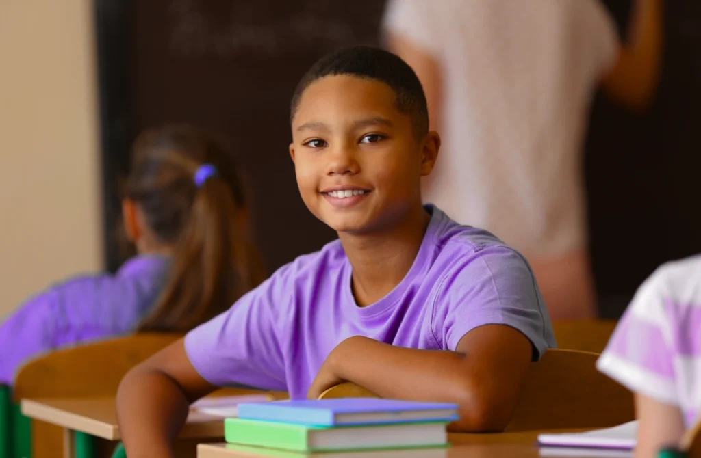 A student wearing a purple shirt sits at a desk in a classroom, smiling, with two books stacked in front of him and classmates in the background.