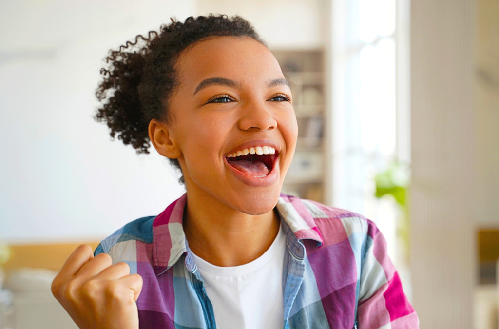 A young person with curly hair, wearing a colorful plaid shirt, smiles and raises a fist in a gesture of excitement or celebration indoors.