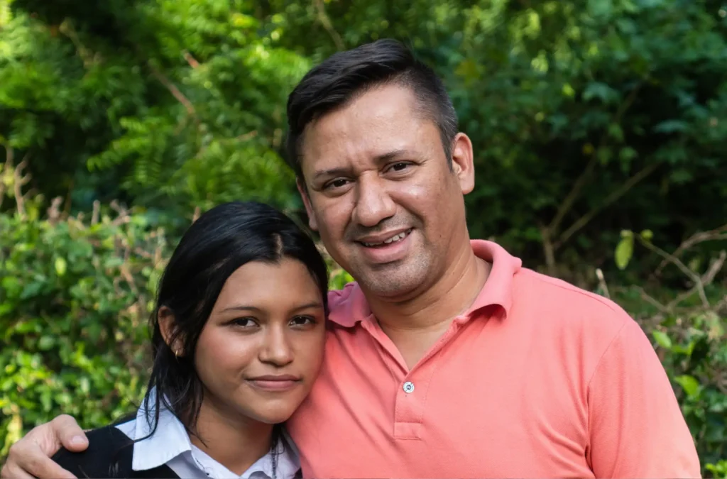 A man in a pink polo shirt stands next to a young woman in a white shirt and black sweater, both smiling outdoors with green foliage in the background.