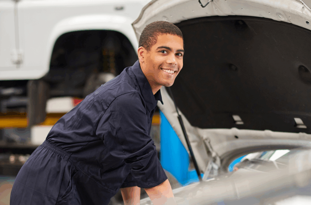 A person in coveralls smiles while working on a car with the hood open in an auto repair shop.