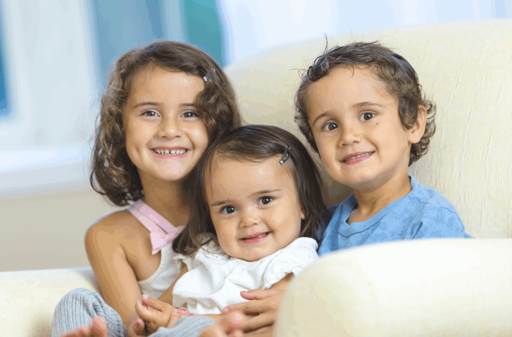 Three young children sit closely together on a light-colored armchair, smiling at the camera.