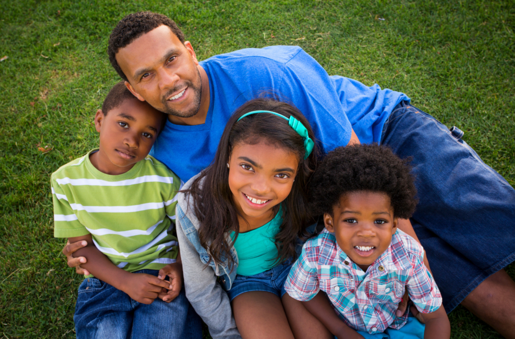 A man sits on grass with three smiling children, two boys and a girl, all looking up at the camera.