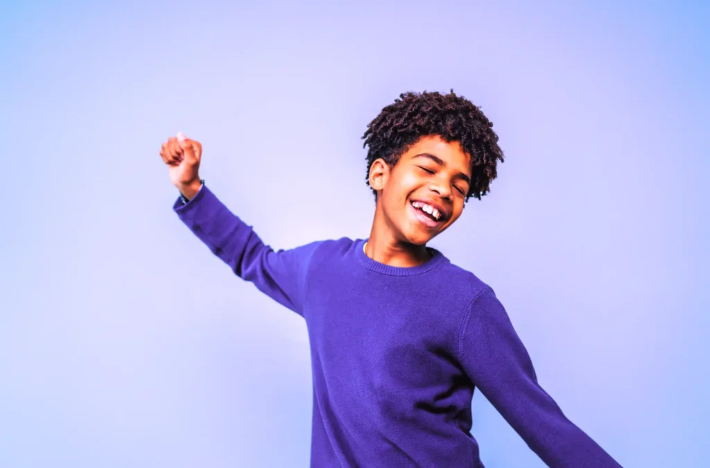 Smiling child in a purple sweater poses with arms raised against a light blue background.