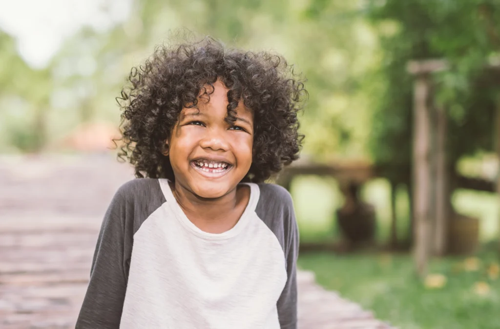 A young child with curly hair wearing a gray and white shirt smiles outdoors, with trees and greenery blurred in the background.