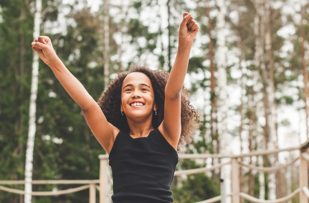 Smiling girl with curly hair in a black tank top raises her arms in the air outdoors, with trees and a wooden fence in the background.