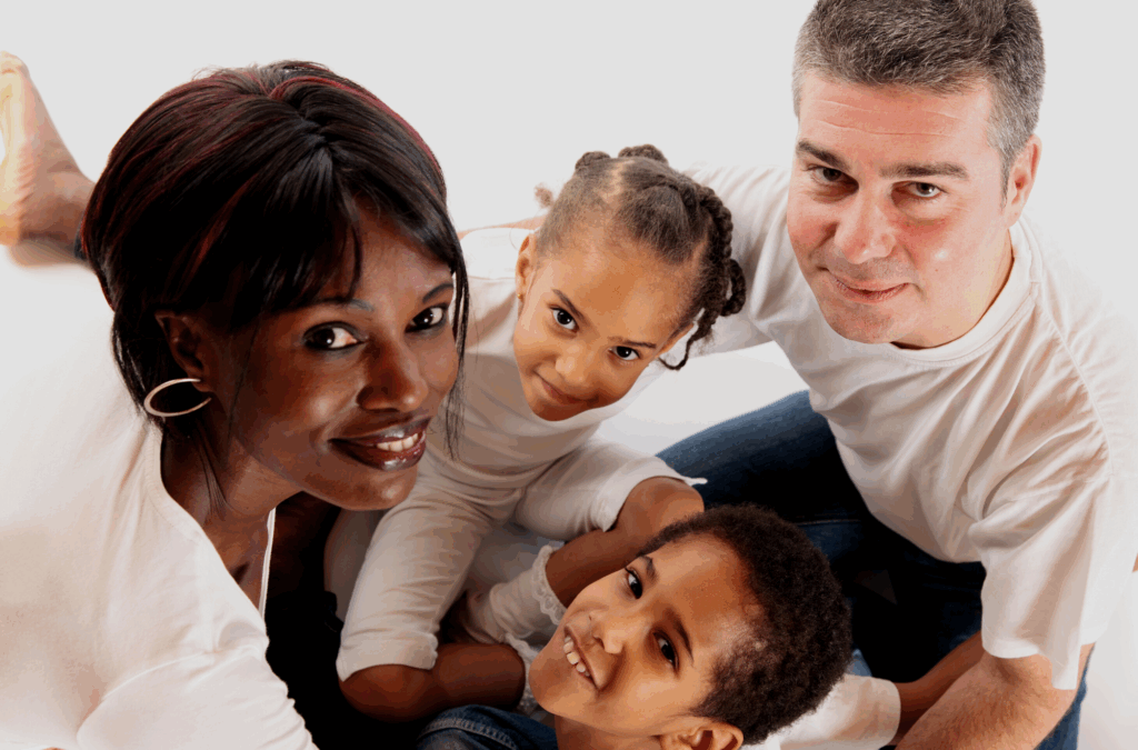 A family of four, two adults and two children, sitting closely together and smiling at the camera, all wearing white shirts against a white background.