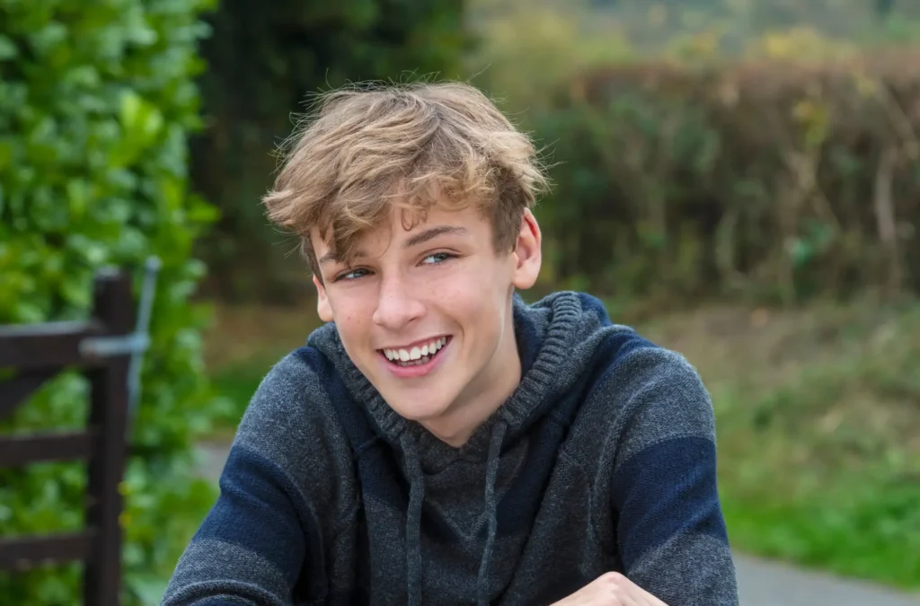 Teen boy with light brown hair wearing a striped hoodie sits outdoors, smiling, with greenery and a wooden bench in the background.