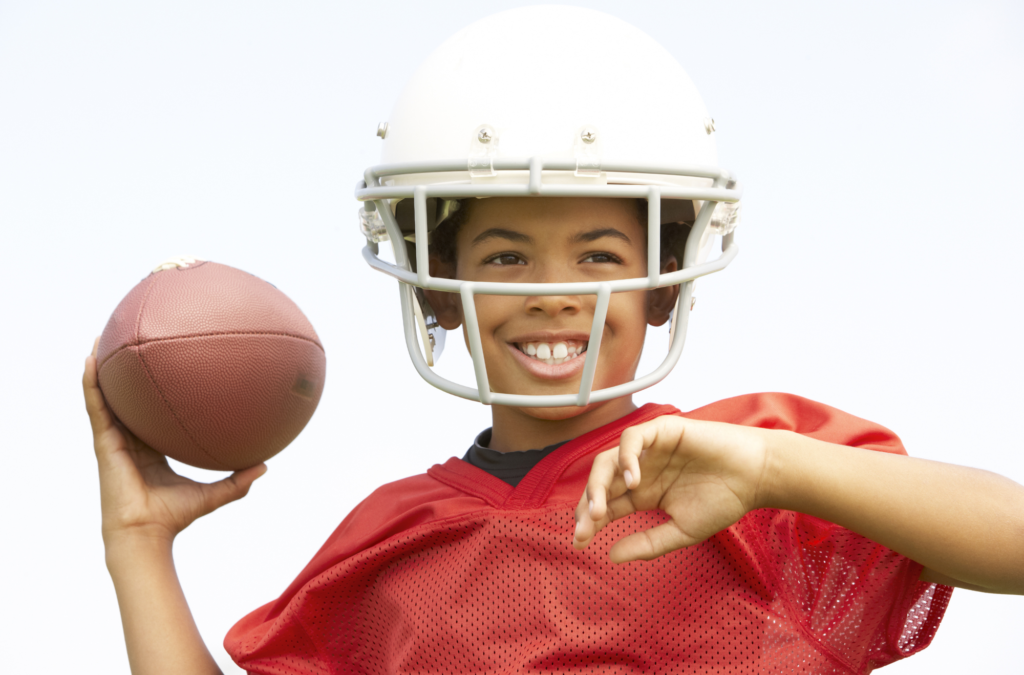 A young boy in a white football helmet and red jersey prepares to throw a football, smiling outdoors under a clear sky.