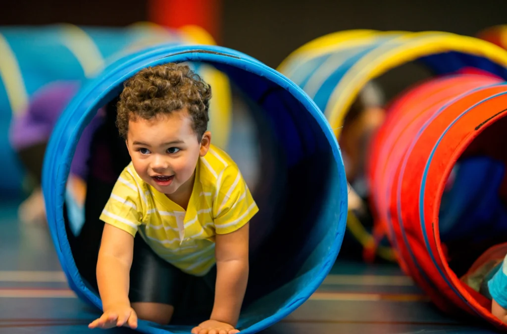 A young child in a yellow striped shirt crawls through a blue play tunnel indoors, smiling and looking forward. Other tunnels are visible in the background.