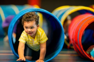 A young child in a yellow striped shirt crawls through a blue play tunnel indoors, smiling and looking forward. Other tunnels are visible in the background.
