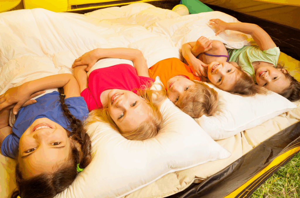 Five children lie on sleeping bags inside a tent, smiling and looking up at the camera, each wearing a different colored shirt.