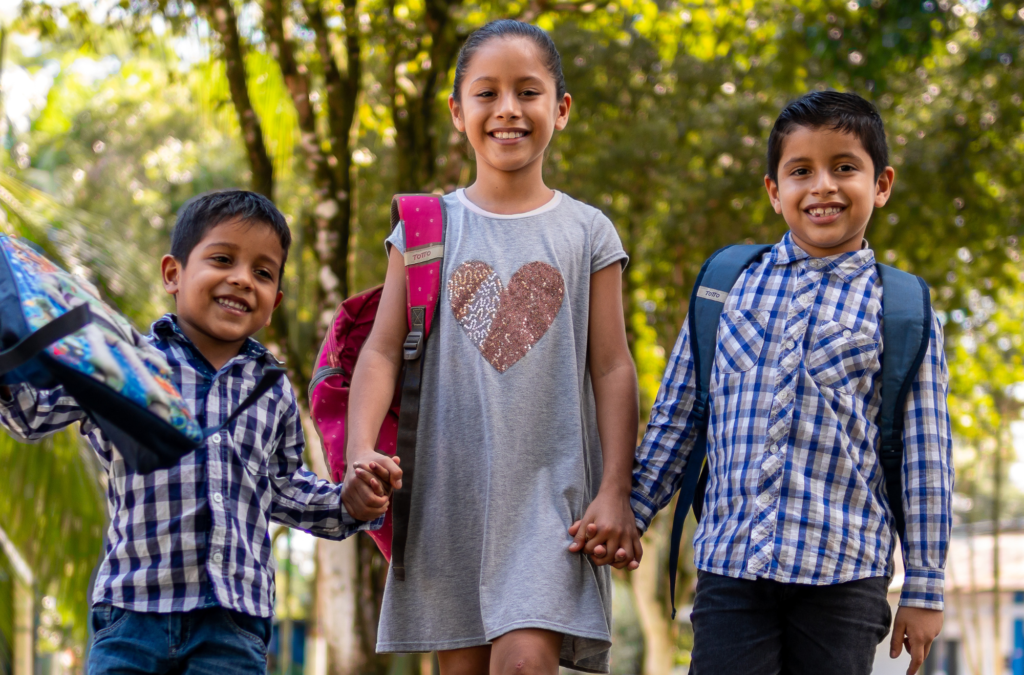 Three smiling children with backpacks, two boys and one girl, holding hands and walking outdoors on a sunny day.