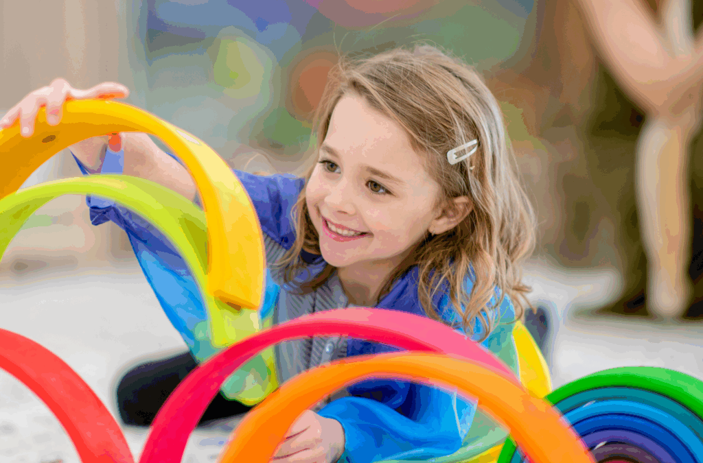 A young child with light brown hair smiles while stacking colorful, curved wooden pieces on the floor indoors.