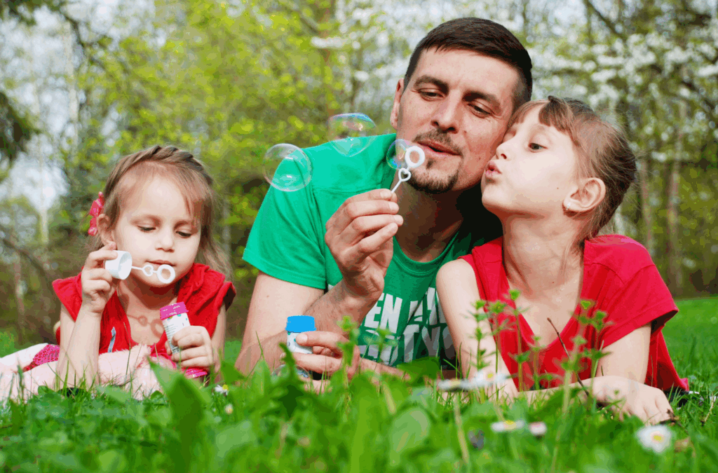 A man and two young girls lie on grass outdoors, blowing soap bubbles together surrounded by greenery and wildflowers.