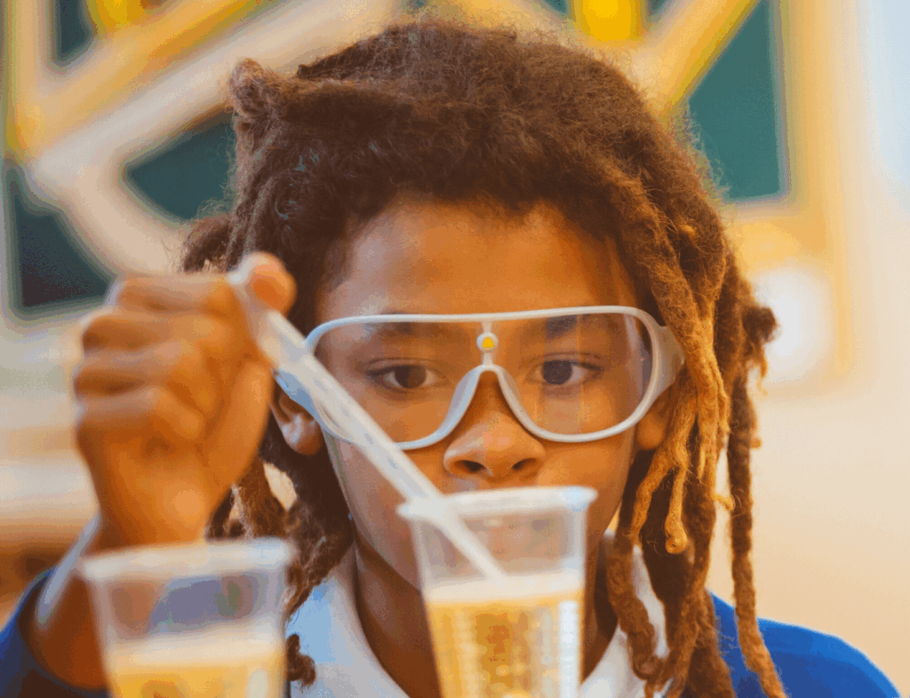 A young student wearing safety goggles uses a pipette to transfer liquid between two clear plastic cups in a classroom or laboratory setting.
