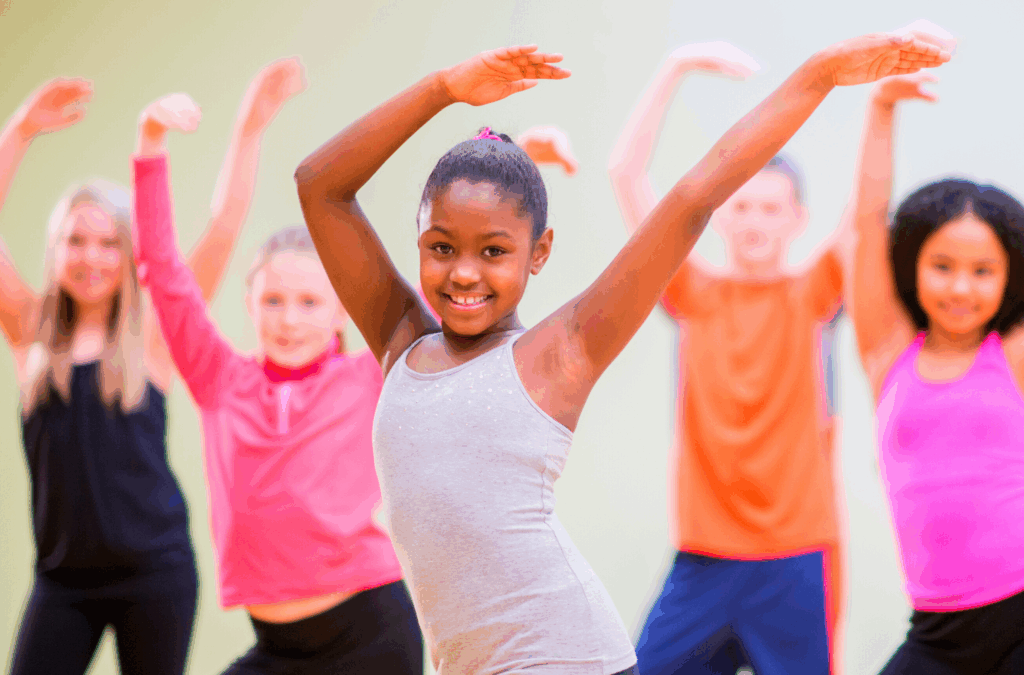 A group of children, led by a girl in a gray tank top, pose with arms raised in a dance or exercise class.