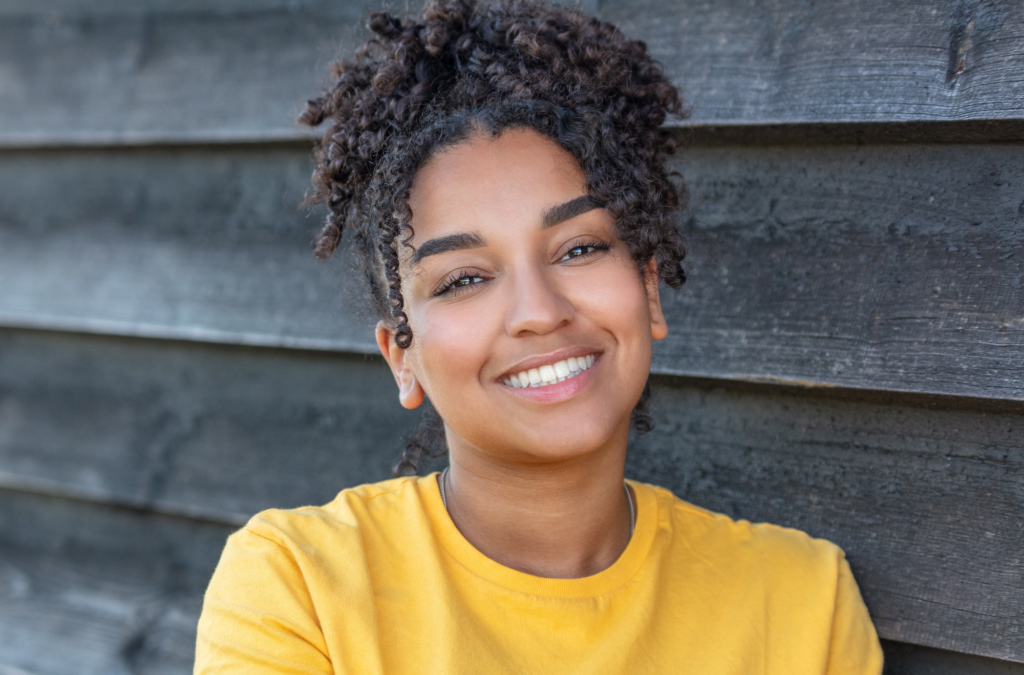 A woman with curly hair, wearing a yellow shirt, smiles while standing in front of a dark wooden wall.