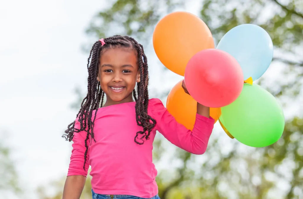 A young girl in a pink shirt smiles while holding four colorful balloons outdoors, with trees visible in the background.