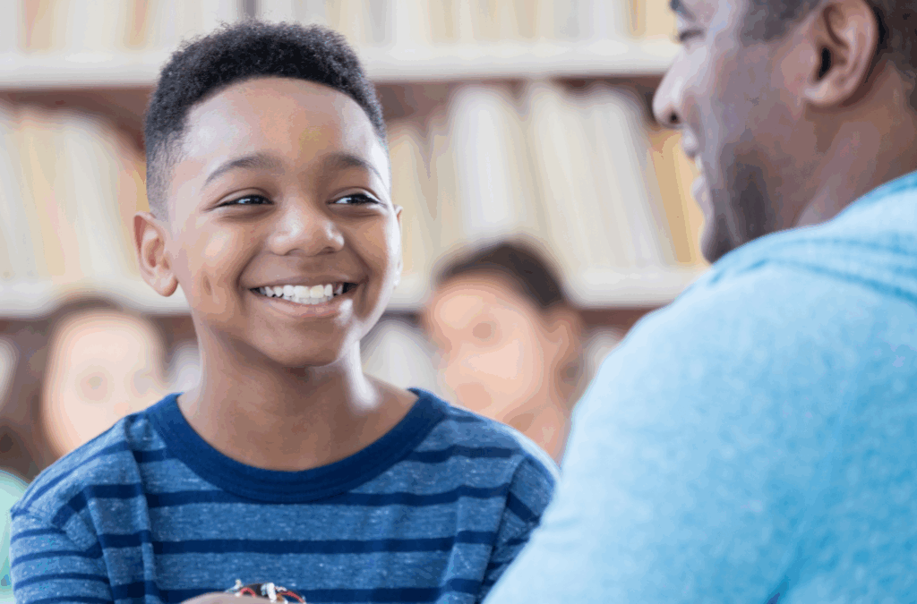 A smiling boy in a blue striped shirt looks at an adult, with blurred bookshelves and people in the background.