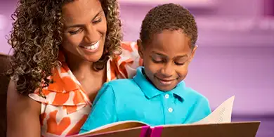 Woman and young boy sit together and smile while looking at an open book, with a blurred purple background.