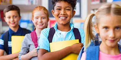 Four schoolchildren with backpacks and folders stand indoors, smiling and looking at the camera.