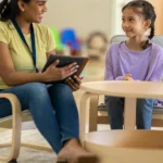 A woman in a yellow shirt sits with a girl in a purple sweater, discussing while holding a tablet, in a cozy room.