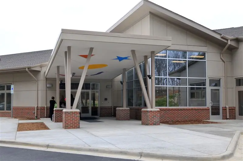 The entrance of a modern building with large windows, brick accents, and a covered walkway decorated with colorful shapes on the ceiling.