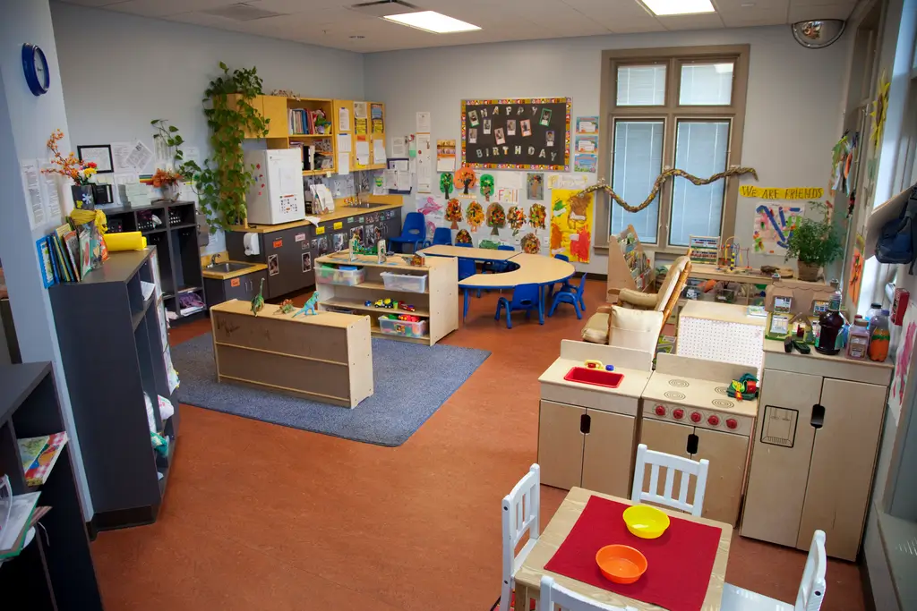 A colorful preschool classroom with small tables, chairs, bookshelves, toys, artwork on walls, and plants, arranged for various activities and play areas.