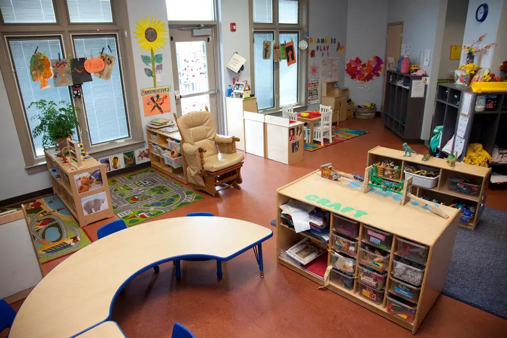 A colorful preschool classroom with small tables, toy shelves, art supplies, children's artwork on the walls, and educational play areas.