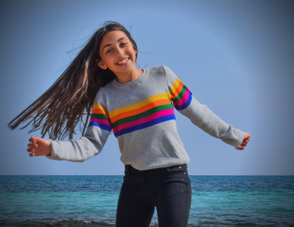 A young girl with long brown hair, wearing a gray sweater with colorful stripes, smiles and poses by the ocean under a clear sky.