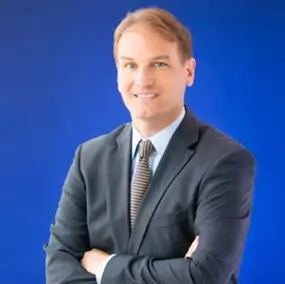 A man in a suit and striped tie stands with arms crossed against a solid blue background, smiling at the camera.