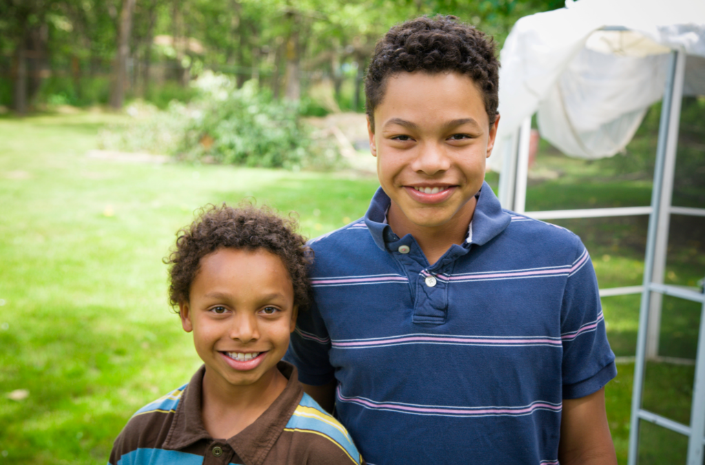 Two boys are standing outside on green grass, smiling at the camera. Trees and part of a white tent or structure are visible in the background.